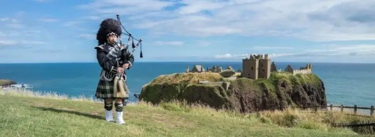 Escocés con vestimenta tradicional tocando la gaita frente al Castillo de Dunnottar en Escocia