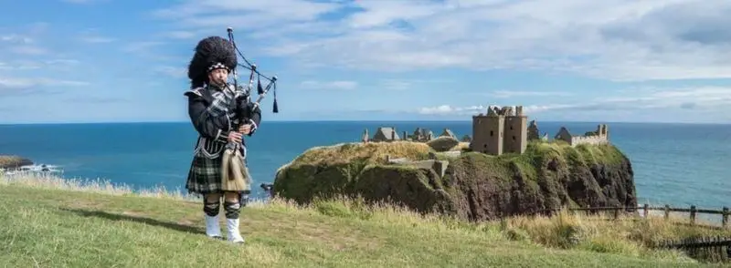Escocés con vestimenta tradicional tocando la gaita frente al Castillo de Dunnottar en Escocia